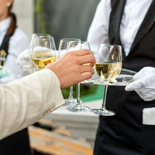 Midsection of professional waiter in uniform serving wine during buffet catering party, festive event or wedding. Full glasses of champagne on tray. Outdoor party catering service, waiter on duty